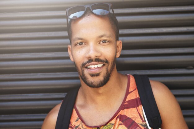 Attrayant étudiant masculin à la peau sombre avec des lunettes de soleil sur la tête à la recherche, ayant une expression de visage joyeuse et insouciante, habillé avec désinvolture, marchant dans la rue après une journée bien remplie à l'université