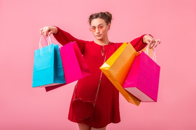 Attrayant heureux émotion drôle femme élégante accro du shopping en robe rouge à la mode tenant des sacs colorés sur le mur rose isolé, vente excité, tendance de la mode printemps été