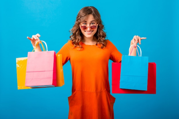 Attrayant heureux émotionnel souriant femme élégante accro du shopping en robe surdimensionnée à la mode orange tenant des sacs sur fond bleu studio isolé