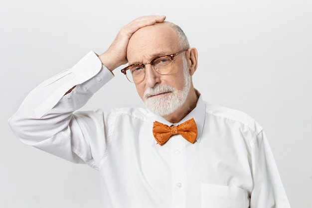 Attrayant homme retraité barbu portant une chemise blanche et un nœud papillon orange tenant la main sur sa tête chauve, se sentant triste parce qu'il a vieilli trop vite. Concept d'âge, de retraite et de maturité