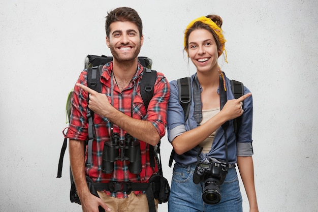 Attrayant jeune couple aventureux amoureux portant des vêtements pratiques, portant des sacs à dos, un appareil photo et des jumelles ayant des regards joyeux, pointant du doigt dans des directions opposées
