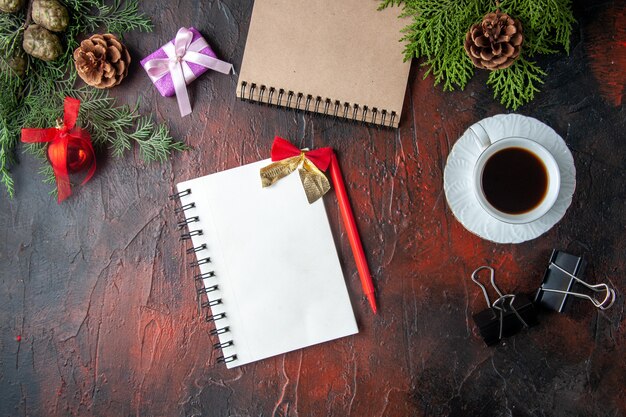 Au-dessus de la vue des branches de sapin, une tasse d'accessoires de décoration de thé noir et un cadeau à côté d'un cahier avec un stylo sur fond sombre