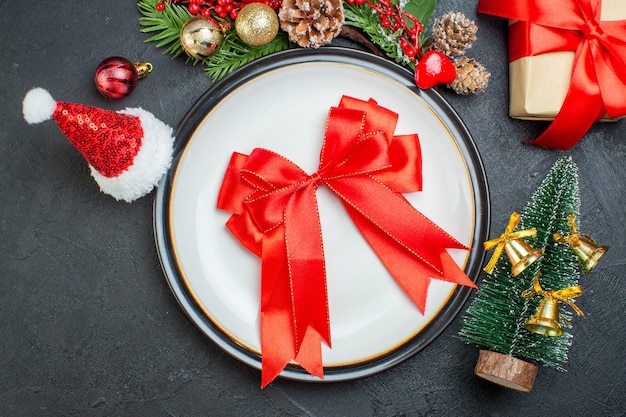 Photo gratuite au-dessus de la vue du ruban rouge en forme d'arc sur une assiette de sapin de noël branches de sapin conifère cône cadeau boîte de santa claus hat sur fond noir