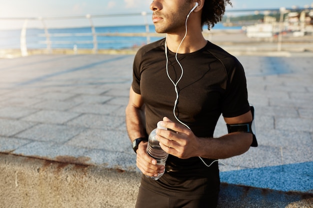 Au milieu du coureur à la peau foncée en tenue de sport noire tenant une bouteille d'eau minérale dans ses mains, en utilisant l'application de musique sur un téléphone mobile pendant un entraînement de jogging derrière la mer.