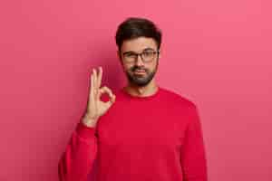 Photo gratuite aucun problème de concept. un homme barbu fait un geste correct, a tout sous contrôle, tout beau geste, porte des lunettes et un pull, pose contre un mur rose, dit que j'ai ça, garantit quelque chose