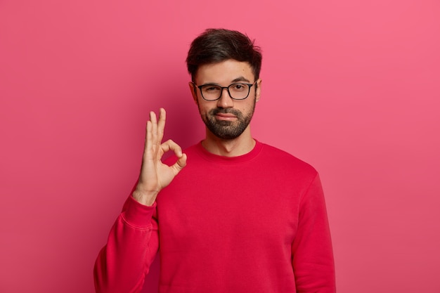 Aucun problème de concept. Un homme barbu fait un geste correct, a tout sous contrôle, tout beau geste, porte des lunettes et un pull, pose contre un mur rose, dit que j'ai ça, garantit quelque chose