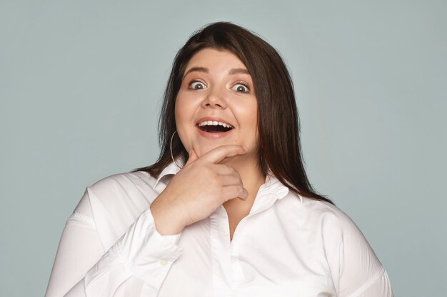 En aucune façon. Portrait de jolie jeune femme potelée en tenue de soirée ouvrant largement la bouche et touchant le menton, exprimant son étonnement tout en écoutant une histoire incroyable. Expressions faciales humaines