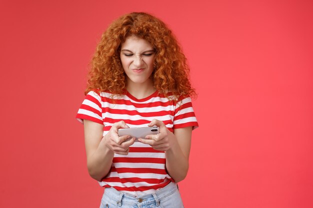 Audacieuse et enthousiaste accro geek belle fille rousse bouclée déteste perdre des batailles jeu de smartphone ...