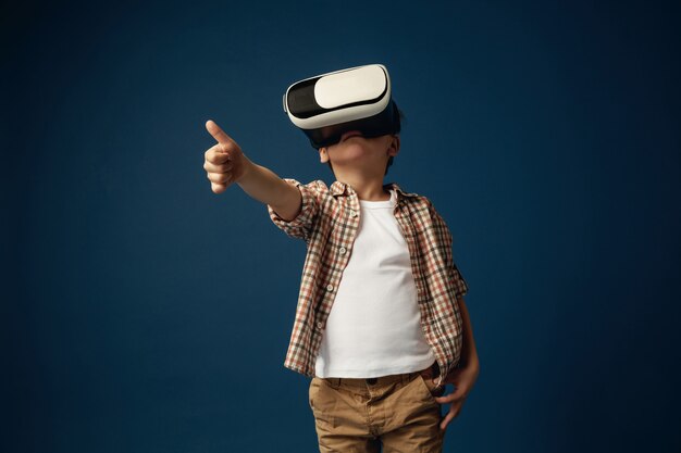 Une autre vision du monde. Petit garçon ou enfant en jeans et chemise avec des lunettes de casque de réalité virtuelle isolés sur fond bleu studio. Concept de technologie de pointe, jeux vidéo, innovation.