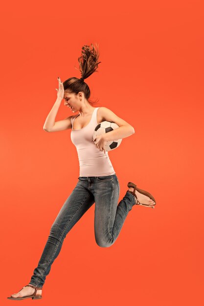 En avant à la victoire.La jeune femme en tant que joueur de football de football sautant et botter le ballon au studio sur un fond rouge. Fan de football et concept de championnat du monde. Concepts d'émotions humaines