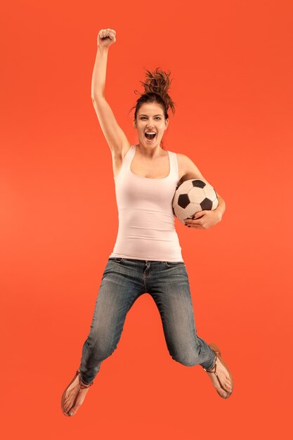 En avant à la victoire.La jeune femme en tant que joueur de football de football sautant et botter le ballon au studio sur un fond rouge. Fan de football et concept de championnat du monde. Concepts d'émotions humaines