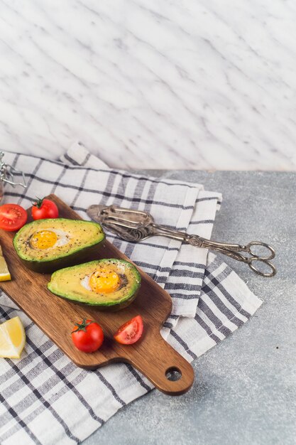 Avocat coupé en deux avec œuf de york; tomates et citron tranche sur une planche à découper sur la serviette avec une pince