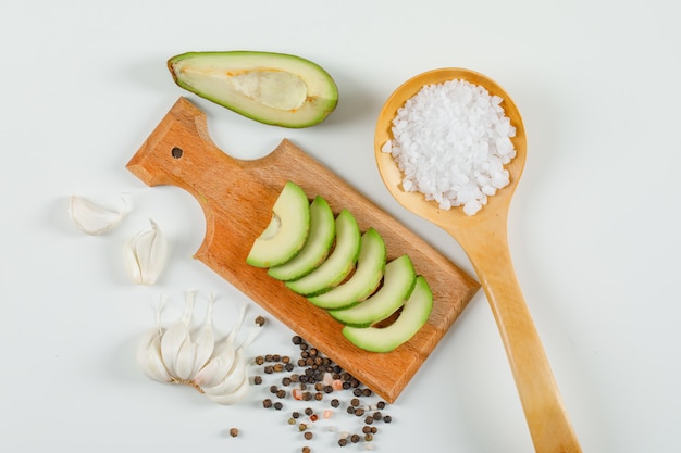 Avocat en tranches dans une planche à découper avec des cristaux de sel dans une cuillère en bois, des gousses d'ail et des grains de poivre vue de dessus sur une surface blanche