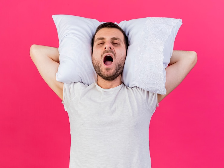 Photo gratuit le bâillement jeune homme malade tenant un oreiller derrière la tête isolé sur rose