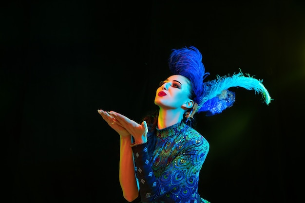 Baisers. Belle jeune femme en carnaval, costume de mascarade élégant avec des plumes sur un mur noir en néon. Copyspace pour l'annonce. Célébration de vacances, danse, mode. Temps de fête, fête.