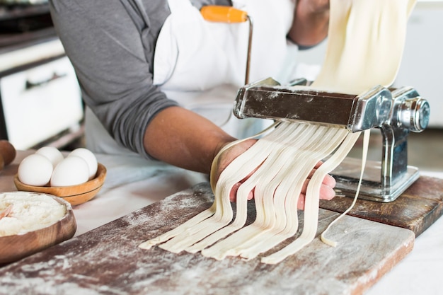 Baker coupe la pâte crue en tagliatelle sur machine à pâtes sur la planche de bois