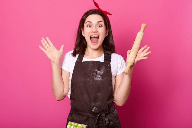 Baker, jeune femme, lève ses mains en tenant un rouleau à pâtisserie, porte un tablier marron, un t-shirt blanc, ouvre largement la bouche. Adorable femme mignonne est de bonne humeur lors de la cuisson de nouveaux plats. Concept de cuisinier.