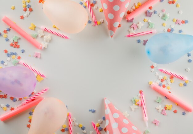 Ballons avec paillettes sucrées, bougies, chapeaux de fête sur un tableau blanc