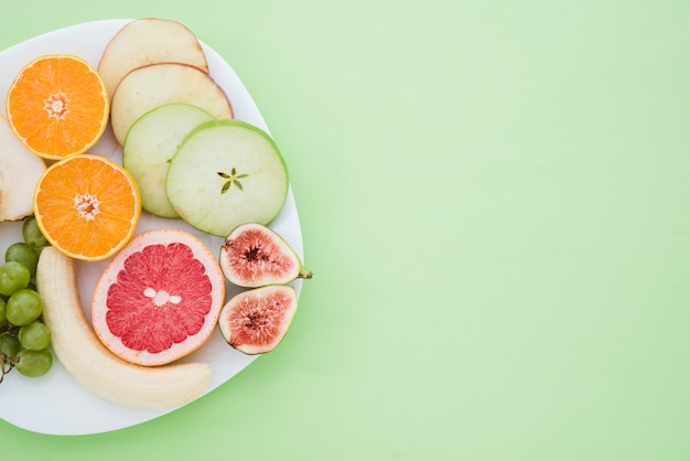 Banane pelée; les raisins; Orange; pamplemousse; figue et tranches de fruits pomme et poire sur une plaque blanche sur le fond vert menthe