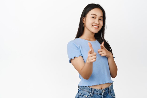 Bang Une fille asiatique souriante pointant du doigt des pistolets à la caméra en vous choisissant de recruter des personnes invitant à un événement debout sur fond blanc