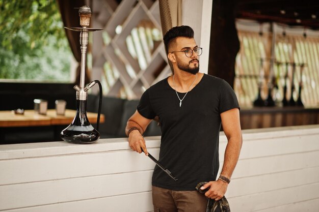 Barbe élégante homme arabe à lunettes et t-shirt noir fumant le narguilé modèle arabe en plein air se reposant