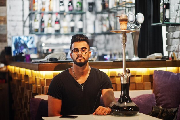 Barbe élégante homme arabe à lunettes et t-shirt noir fumer narguilé bar intérieur modèle arabe se reposer