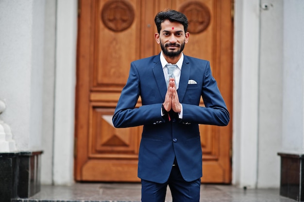 Photo gratuite barbe élégante homme indien avec bindi sur le front porter sur costume bleu posé en plein air contre la porte du bâtiment et montrer le signe des mains namaste