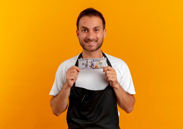 Barber man in apron holding cash à l'avant avec le sourire sur le visage debout sur un mur orange