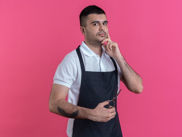 Barber man in apron holding ciseaux looking at camera perplexe debout sur fond rose