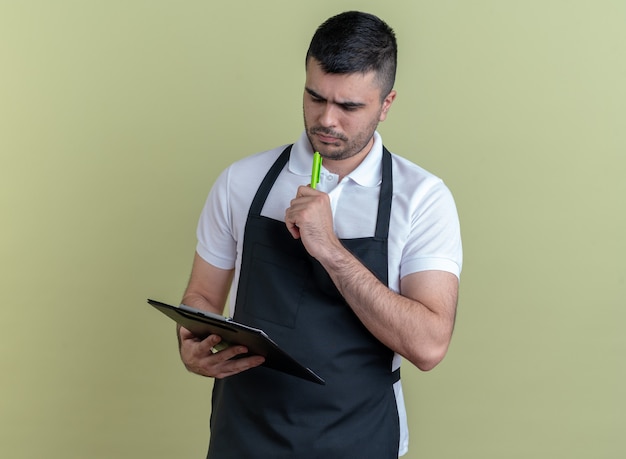 Barber man in apron holding clipbord et stylo le regardant avec un visage sérieux debout sur fond vert