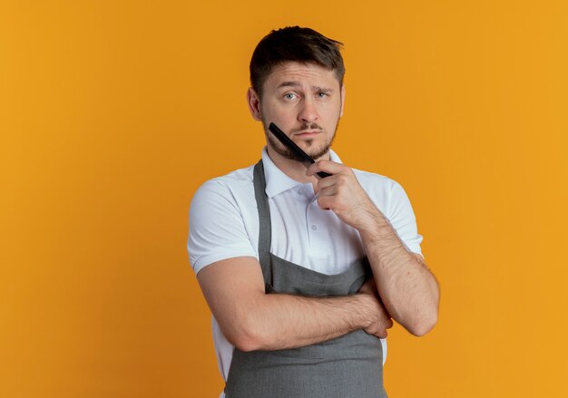 Barber man in apron holding comb lookign at camera avec visage sérieux debout sur fond orange