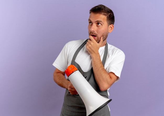 Barber man in apron holding holding megaphone à côté perplexe debout sur le mur violet