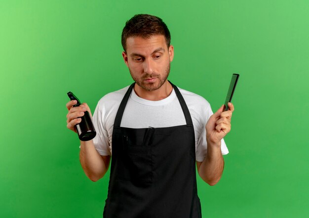 Barber man in apron holding spray et peigne à côté perplexe debout sur mur vert