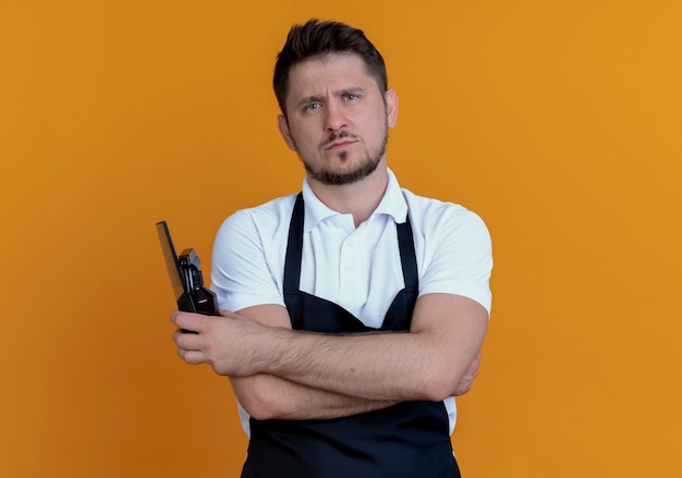 Barber man in apron holding spray et peigne regardant la caméra avec un visage sérieux debout sur fond orange