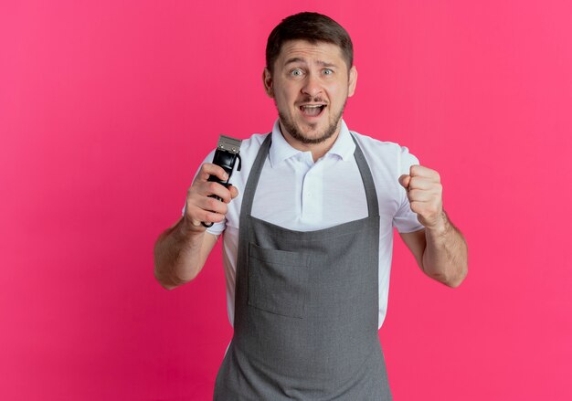 Barber man in apron holding tondeuse à barbe regardant la caméra serrant le poing heureux et excité debout sur fond rose