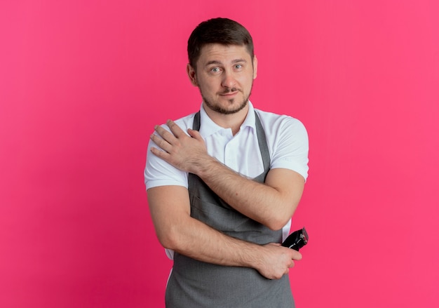 Barber man in apron holding trimmer looking at camera avec une expression confiante sérieuse debout sur fond rose