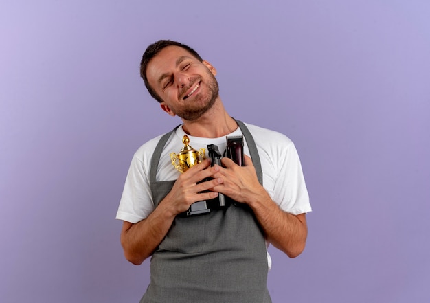 Barber Man In Apron Holding Trophy Et Machine De Coupe De Cheveux Souriant Sentiment Reconnaissant Avec Les Yeux Fermés Debout Sur Le Mur Violet