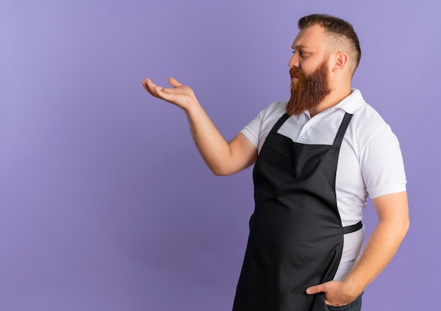 Barbier barbu professionnel en tablier présentant l'espace de copie avec le bras de sa main debout sur le mur violet