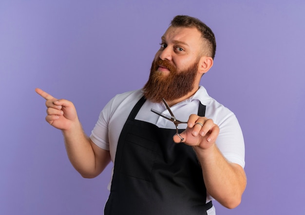 Barbier barbu professionnel en tablier tenant des ciseaux à la confiance en pointant avec le doigt sur le côté debout sur le mur violet