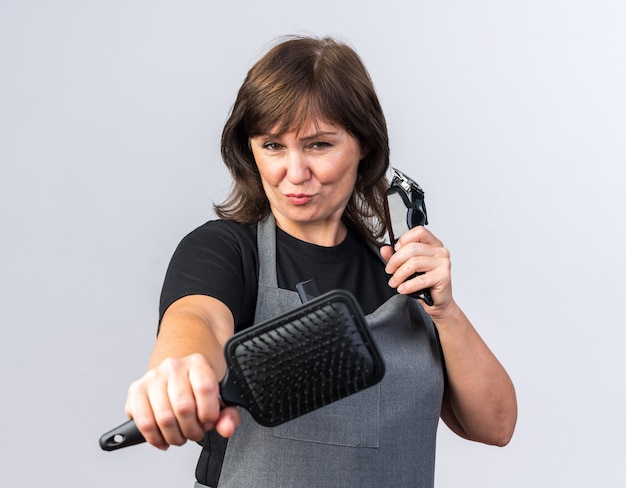 Barbier femme adulte confiant en uniforme tenant une tondeuse à cheveux et un peigne isolé sur un mur blanc avec espace de copie