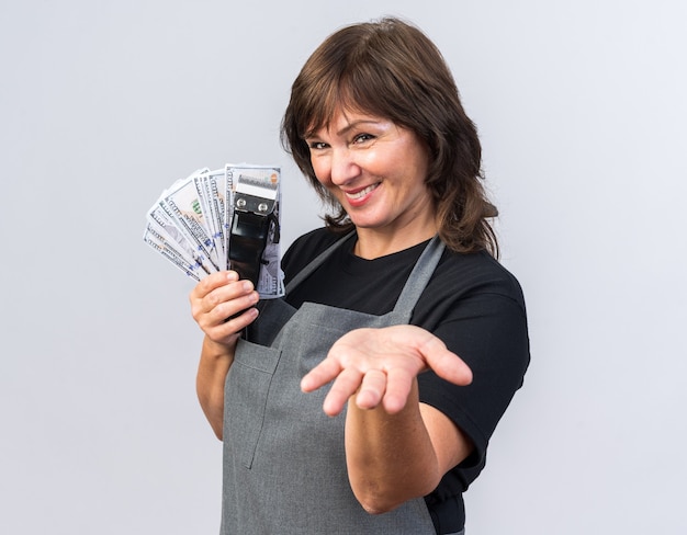 Photo gratuite barbier femme adulte souriante en uniforme tenant une tondeuse à cheveux avec de l'argent et gardant la main ouverte isolée sur un mur blanc avec espace de copie