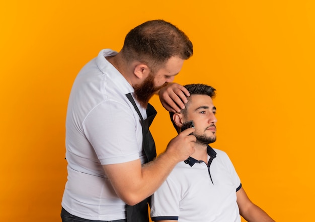 Barbier professionnel barbu en tablier faisant coupe de cheveux avec machine à raser à un jeune homme debout sur un mur orange
