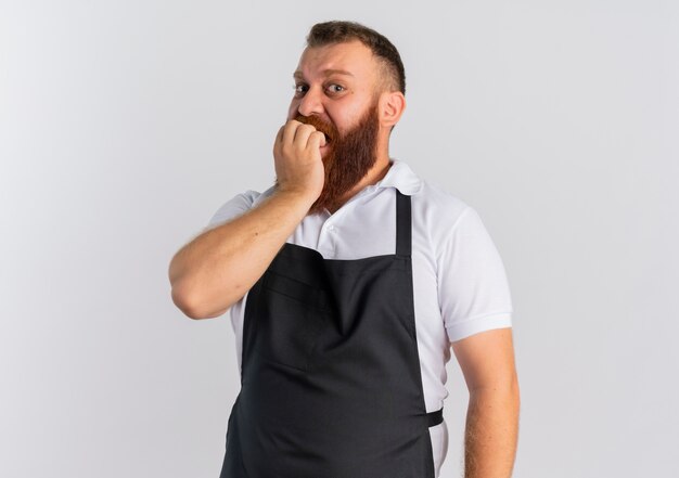 Barbier professionnel barbu en tablier a souligné et nerveux mordant les ongles debout sur un mur blanc