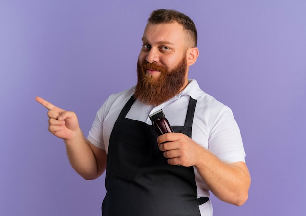 Barbier professionnel barbu en tablier tenant la machine de coupe de cheveux pointant avec le doigt sur le côté souriant debout sur le mur violet