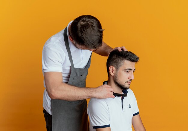 Barbier en tablier barbe de rasage avec rasoir de client satisfait sur mur orange