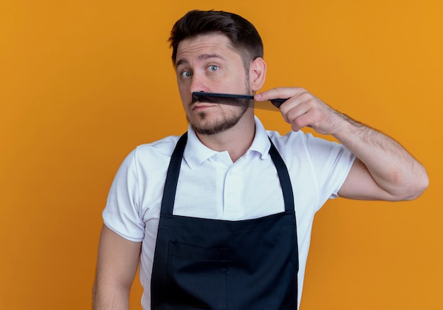 Barbier en tablier peignant sa barbe regardant la caméra debout sur fond orange