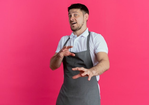 Barbier en tablier regardant la caméra avec une expression dégoûtée faisant un geste de défense avec les mains debout sur fond rose