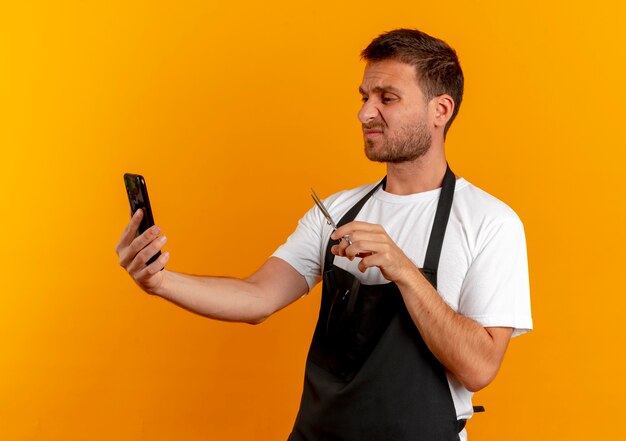 Barbier en tablier regardant l'écran de son téléphone portable tenant des ciseaux à la mécontentement debout sur le mur orange