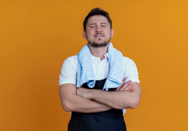 Barbier en tablier avec une serviette autour du cou souriant confiant avec les bras croisés debout sur le mur orange