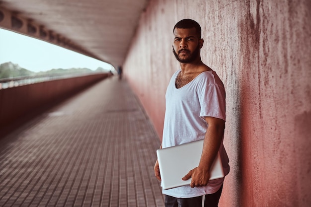 Un barbu afro-américain vêtu d'une chemise blanche et d'un short de sport tient sur un ordinateur portable tout en se tenant sur une passerelle sous un pont.